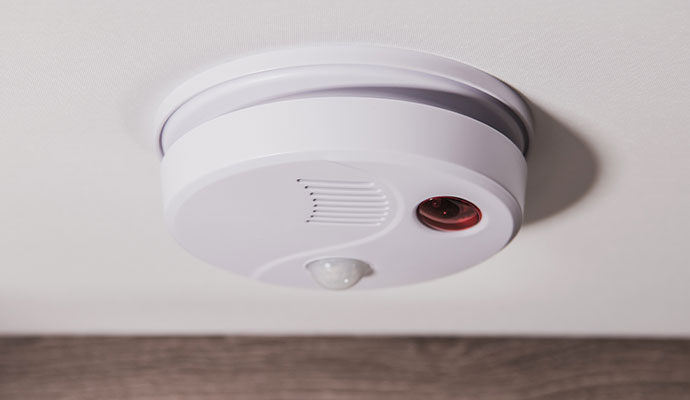 A white ceiling-mounted alarm device featuring a motion sensor lens, speaker vents, and a red indicator light