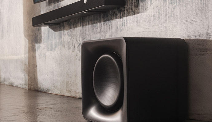 A close-up side profile of a compact black KEF subwoofer on a concrete floor