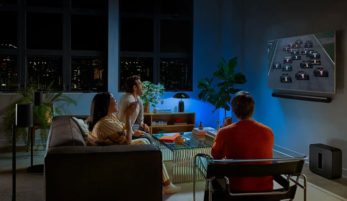 A group of friends in a dimly lit modern living room watching a car race on a large TV equipped with a Sonos soundbar