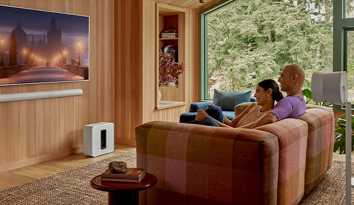 A couple relaxing on a plaid sofa in a wood-paneled living room featuring a white Sonos Arc soundbar