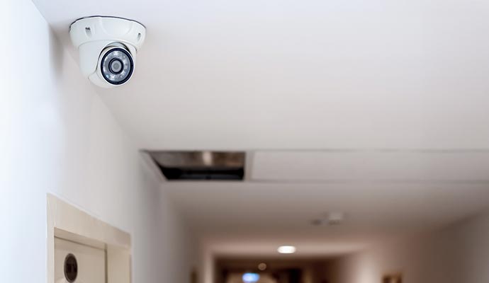A white dome-shaped CCTV security camera is mounted on a ceiling in a long, well-lit indoor corridor.