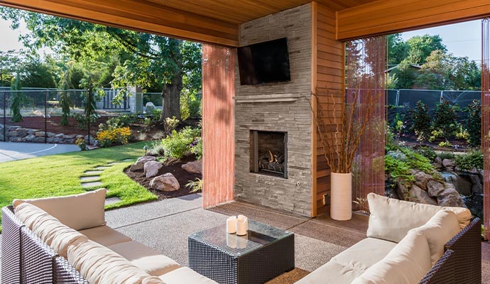 Modern outdoor living area featuring a stone fireplace, wall-mounted TV, and wicker sofas overlooking a lush backyard